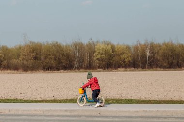 Kırmızı ceketli ve yeşil şapkalı bir çocuk bir tarlanın yanındaki asfalt yolda mavi dengeli bir bisiklete biniyor. Çocuk kameradan uzağa bakıyor ve arkaplandaki alan kahverengi. Güneşli bir gün.