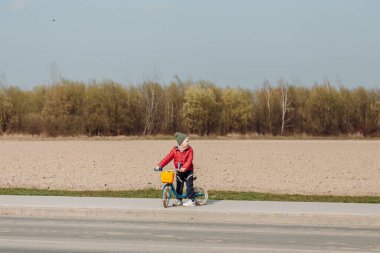 Kırmızı ceketli ve yeşil şapkalı bir çocuk bir tarlanın yanındaki asfalt yolda mavi dengeli bir bisiklete biniyor. Çocuk kameradan uzağa bakıyor ve arkaplandaki alan kahverengi. Güneşli bir gün.