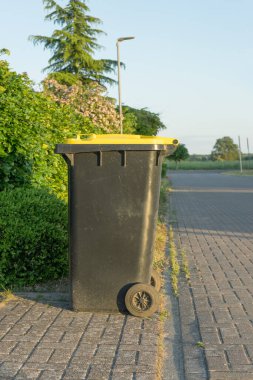 Yol kenarında sarı kapaklı siyah çöp tenekesi.