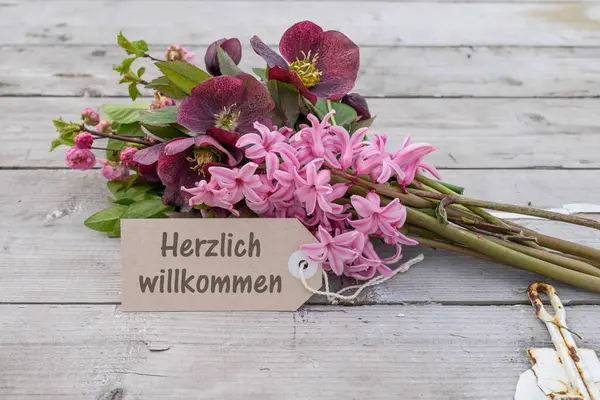 ドイツ語のテキストが付いているピンクのヒヤシンス クリスマスのバラおよびカードが付いている花束 暖かく歓迎 ストック画像