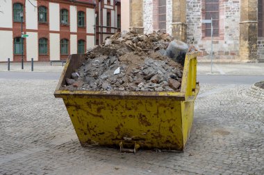 İnşaat atıklarıyla dolu sarı metal bir konteynır tarihi binaların önündeki meydanda duruyor.