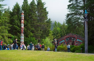 Saxman, AK, ABD - 27 Haziran 2024: 25 kutuptan ikisi yakınında Ketchikan yakınlarındaki Saxman Totem parkında bir tur grubu toplandı. Arka planda Kunduz Klanı Evi duruyor..