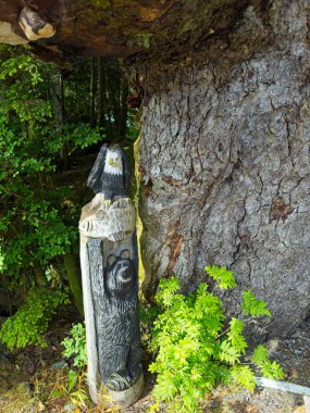 Ketchikan, AK, USA - June 27, 2024: A small wooden totem in depicting a bear, fish, and bald eagle in a natural style stands at the base of a large tree in a nature sanctuary near Ketchikan. clipart