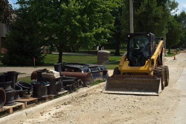 Twinsburg, OH, USA - 18 Temmuz 2024: Bir patinaj dümeni yükleyicisi ve çeşitli su yenileme parçaları yaz inşaat projesi sırasında bir iskan caddesinin yanında ve yanında oturur.