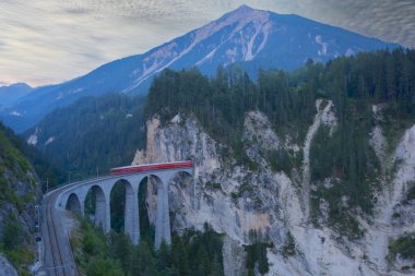A local train of Rhaetian Railway coming out of the tunnel in a cliff crossing famous Landwasser Viaduct over a deep gorge with fall colors on the rocky mountainside in Filisur, Grisons, Switzerland