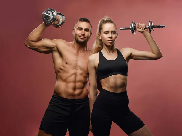 Fitness male and female models with equipment at pink background. Sporty couple with dumbbells in studio. Shirtless muscular man and slim girl with six pack abs posing with gym stuff