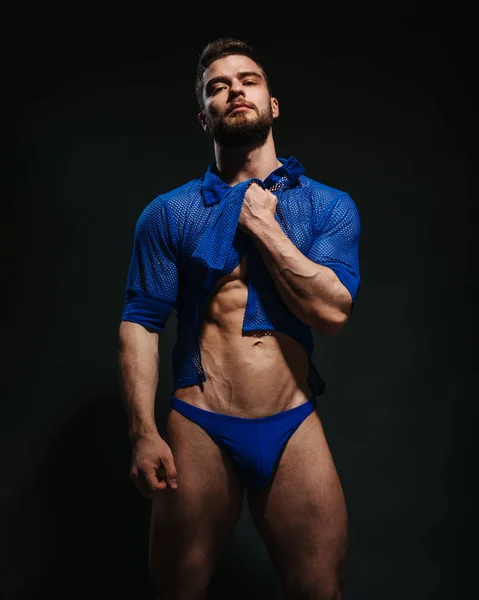 Sexy muscled male model in blue shirt and blue swimwear on black background. Bearded man taking his clothes up. Fashion guy in beachwear in studio. Male fitness model in stylish outlook.