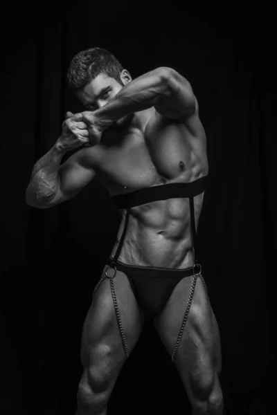 Handsome muscular man in harness with chains and black briefs. Six pack abs model on black background. Sexy male body in black fetish wear. Black and white body shot of muscular man.