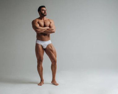 Beyaz iç çamaşırlı seksi kaslı adam stüdyoda poz veriyor. Gri arka planda duran yakışıklı vücut geliştirici. Büyük erkek model kaslarını gösteriyor..