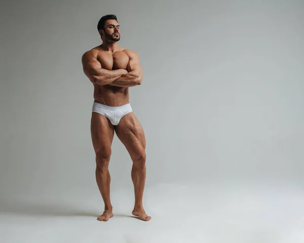 stock image Sexy muscular man in white underwear posing in studio. Handsome bodybuilder standing on grey background. Big male model showing his muscles.