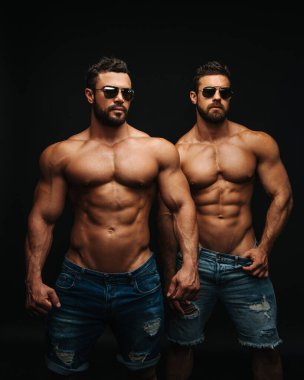 Two shirtless hunks at black background. Fitness models couple in jeans and sunglasses. Two muscular handsome men with six pack abs in studio.