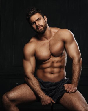 Brutal sexy man with six pack abs in towel at black background. Handsome fitness male model sitting in studio. Bearded muscled guy covering with towel in studio.