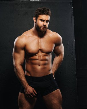 Naked man in black underwear on black background. Sexy muscled guy with six pack abs posing in studio. Bearded brutal macho looking at camera. Male fitness model portrait.