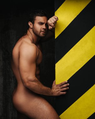 Naked man near wall. Male fitness model posing without wear in studio. Handsome guy showing his muscular body on black background.