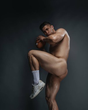 Naked handsome guy with american football ball. Naked American football player is preparing for throw in studio. Bodybuilder playing sports.