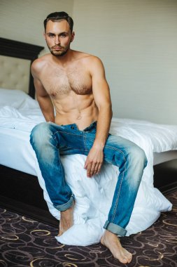 Sakallı, kot pantolonlu, yatakta oturan üstsüz bir adam. Otel odasında karın kası olan uzun saçlı bir erkek model. Kıllı göğüslü kaslı seksi adam..