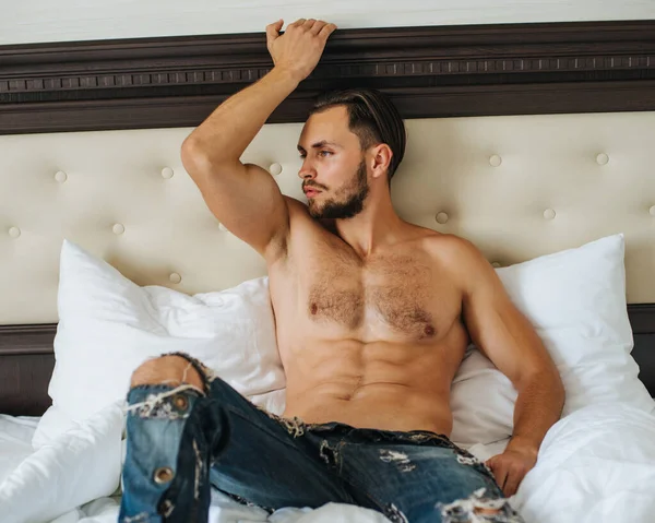 Long hair male model in blue jeans sitting on bed. Shirtless man with six pack abs in hotel room. Naked sexy guy posing on white sheet.