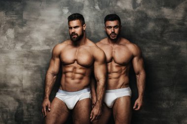 Duvarın yanında duran iki çıplak adam. Stüdyoda beyaz iç çamaşırlı iki fitness mankeni poz veriyor. Birkaç kaslı adam kameraya bakıyor..
