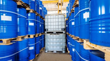 Mavi metal fıçılar ve büyük plastik kaplar sağlam paletler üzerinde dinleniyor, hareketli bir deponun organize kaosunun yankıları, titreşen floresan lambaların altında harekete geçmeye hazır.