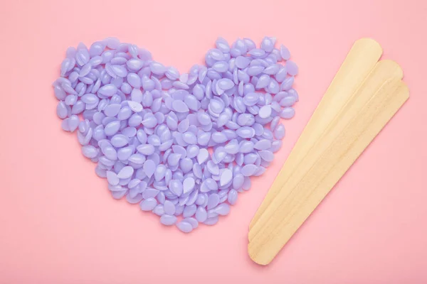 Wooden sticks for wax and spa with wax granules in the shape of a purple heart on a pink background. Top view.