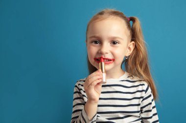 Mutlu küçük kız mavi arka planda dudaklarına ruj sürüyor. Biraz moda. Makyaj yapan küçük kız. Üst görünüm