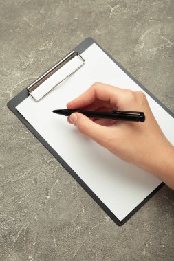Bir kadın eli, üzerinde gri beton arka plan yazılı bir kalem ve siyah bir pano tutuyor. Üst görünüm