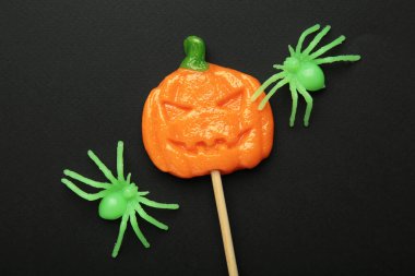 Cadılar Bayramı lolipopları balkabağı şeklinde ve siyah arka planda dekoratif örümcek var. Mevsimsel kutlamalar için bayram ziyafetleri. Üst görünüm