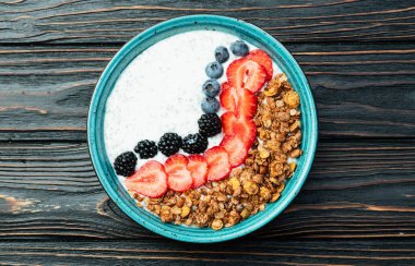 Sağlıklı kahvaltı. Çilekli yoğurt (çilek, ahududu, yabanmersini, böğürtlen), granola ve chia tohumları. Kırsal arkaplanda üst görünüm