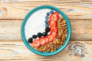 Sağlıklı kahvaltı. Çilekli yoğurt (çilek, ahududu, yabanmersini, böğürtlen), granola ve chia tohumları. Kırsal arkaplanda üst görünüm