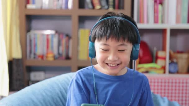 青いヘッドフォンの接続中の幸せな若いアジアの少年は 自宅でベッドの上の音楽を聞いている間 スマートフォンを使用して笑顔幸せな — ストック動画