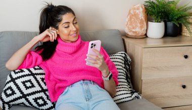 Güzel, mutlu, pembe kazaklı bir kadın evde oturuyor ve cep telefonunu kullanıyor.