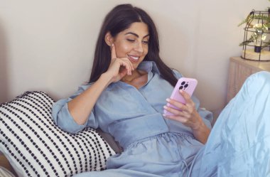 Güzel, mutlu, pembe kazaklı bir kadın evde oturuyor ve cep telefonu kullanıyor.