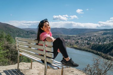 Güzel, gülümseyen bir kadın, mavi bir gölün üzerinde, muhteşem manzaralı bir bankta oturuyor. 