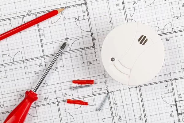 Smoke detector or fire alarm sensor on white architectural plans background with tools and screws, house safety or security concept, flat lay top view from above