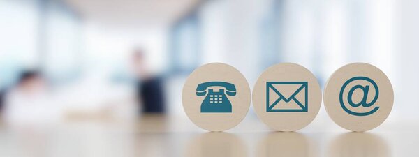 Wooden round telephone, envelope letter and e-mail symbols in a row on wooden table with a blurred office in the background, contact us symbols or banner with copy space, 3D illustration