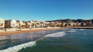 Kıyı şehrinin panoramik insansız hava aracı görüntüsü güneşli bir günde görülen eski Sitges kasabası. Akdeniz 'de dalgalanan deniz dalgalarına yakın çekim. Barcelona ili, Katalonya, İspanya. Film tanıtımı. 4K video