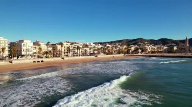 Kıyı şehrinin panoramik insansız hava aracı görüntüsü güneşli bir günde görülen eski Sitges kasabası. Akdeniz 'de dalgalanan deniz dalgalarına yakın çekim. Barcelona ili, Katalonya, İspanya. Film tanıtımı. 4K video