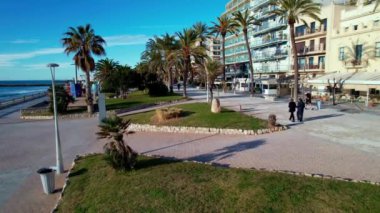 Kıyı şehrinin panoramik insansız hava aracı görüntüsü güneşli bir günde görülen eski Sitges kasabası. Akdeniz 'de dalgalanan deniz dalgalarına yakın çekim. İnsanlar set boyunca yürüyorlar. İspanya. Film tanıtımı. 4K video