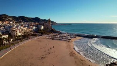 İspanya 'nın Katalonya kenti Barselona yakınlarındaki Sitges kasabasında güneşli bir günde Sant Bartomeu i Santa Tecla de Sitges kıyı köyünün panoramik manzarası. Plajlarıyla ünlüdür. Film tanıtımı. 4K video