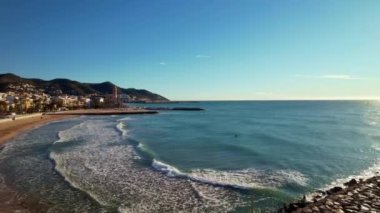 Kıyı şehrinin panoramik insansız hava aracı görüntüsü güneşli bir günde görülen eski Sitges kasabası. Akdeniz 'de dalgalanan deniz dalgalarına yakın çekim. Barcelona ili, Katalonya, İspanya. Film tanıtımı. 4K video
