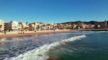 Kıyı şehrinin panoramik insansız hava aracı görüntüsü güneşli bir günde görülen eski Sitges kasabası. Akdeniz 'de dalgalanan deniz dalgalarına yakın çekim. Barcelona ili, Katalonya, İspanya. Film tanıtımı. 4K video