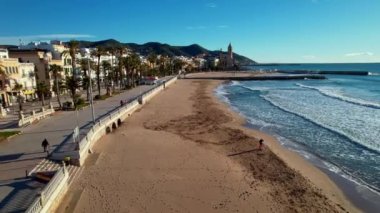 Kıyı şehrinin panoramik insansız hava aracı görüntüsü güneşli bir günde görülen eski Sitges kasabası. Akdeniz 'de dalgalanan deniz dalgalarına yakın çekim. İnsanlar set boyunca yürüyorlar. İspanya. Film tanıtımı. 4K video