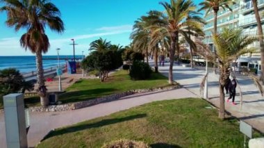 Sitges Akdeniz 'deki sahil ve otellerden oluşan palmiye ağaçlarıyla çevrili güzel plajların manzarası. Avuçların arasında uçan bir dron. İnsanlar set boyunca yürüyorlar. Yaz tatili kavramı. 4K