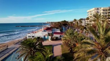 Sitges Akdeniz 'deki sahil ve otellerden oluşan palmiye ağaçlarıyla çevrili güzel plajların manzarası. Avuçların arasında uçan bir dron. İnsanlar set boyunca yürüyorlar. İspanya. 4K video