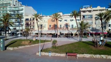 Sitges 'de palmiye ağaçları, plaj ve otellerle çevrili güzel plajların manzarası. Avuçların arasında uçan bir dron. İnsanlar set boyunca yürüyorlar. Yaz tatili kavramı. İspanya. 4K video