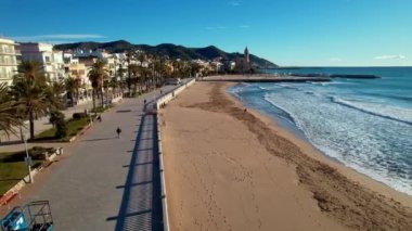 İspanya 'nın Katalonya kenti Barselona yakınlarındaki Sitges kasabasında güneşli bir günde Sant Bartomeu i Santa Tecla de Sitges kıyı köyünün panoramik manzarası. İnsanlar set boyunca yürüyorlar. Film tanıtımı. 4K