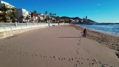 Sant Bartomeu i Santa Tecla de Sitges kıyı köyünün panoramik hava aracı görüntüsü. Köpekli bir adam dolgu seti boyunca yürüyor. Deniz dalgalarının yaz manzarası kıyıya vurur, güzel plajlar, beyaz balyalar