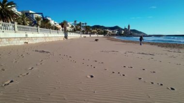 Sant Bartomeu i Santa Tecla de Sitges kıyı köyünün panoramik hava aracı görüntüsü. Köpekli bir adam dolgu seti boyunca yürüyor. Deniz dalgalarının yaz manzarası kıyıya vurur, güzel plajlar, beyaz balyalar