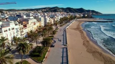 Sant Bartomeu i Santa Tecla de Sitges kıyı köyünün panoramik hava aracı görüntüsü. İnsanlar, denize bakan klasik beyaz balyoz ve boş teraslarla dolgu seti boyunca yürüyorlar. 4K