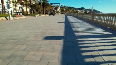 Sant Bartomeu i Santa Tecla de Sitges kıyı köyünün panoramik hava aracı görüntüsü. İnsanlar, denize bakan klasik beyaz balyoz ve boş teraslarla dolgu seti boyunca yürüyorlar. 4K